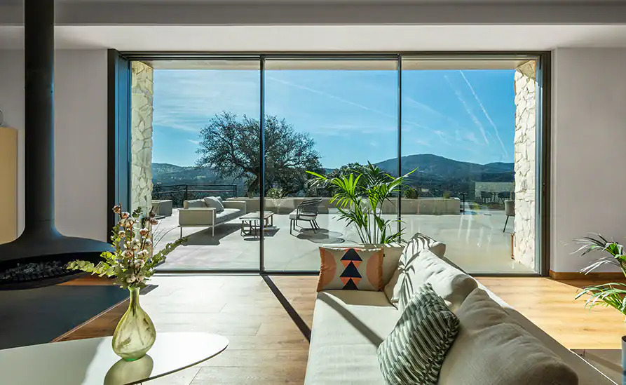 Sofa placed directly beside a floor-to-ceiling aluminium sliding door, with sunlight falling across the floor where occupants sit near the glass